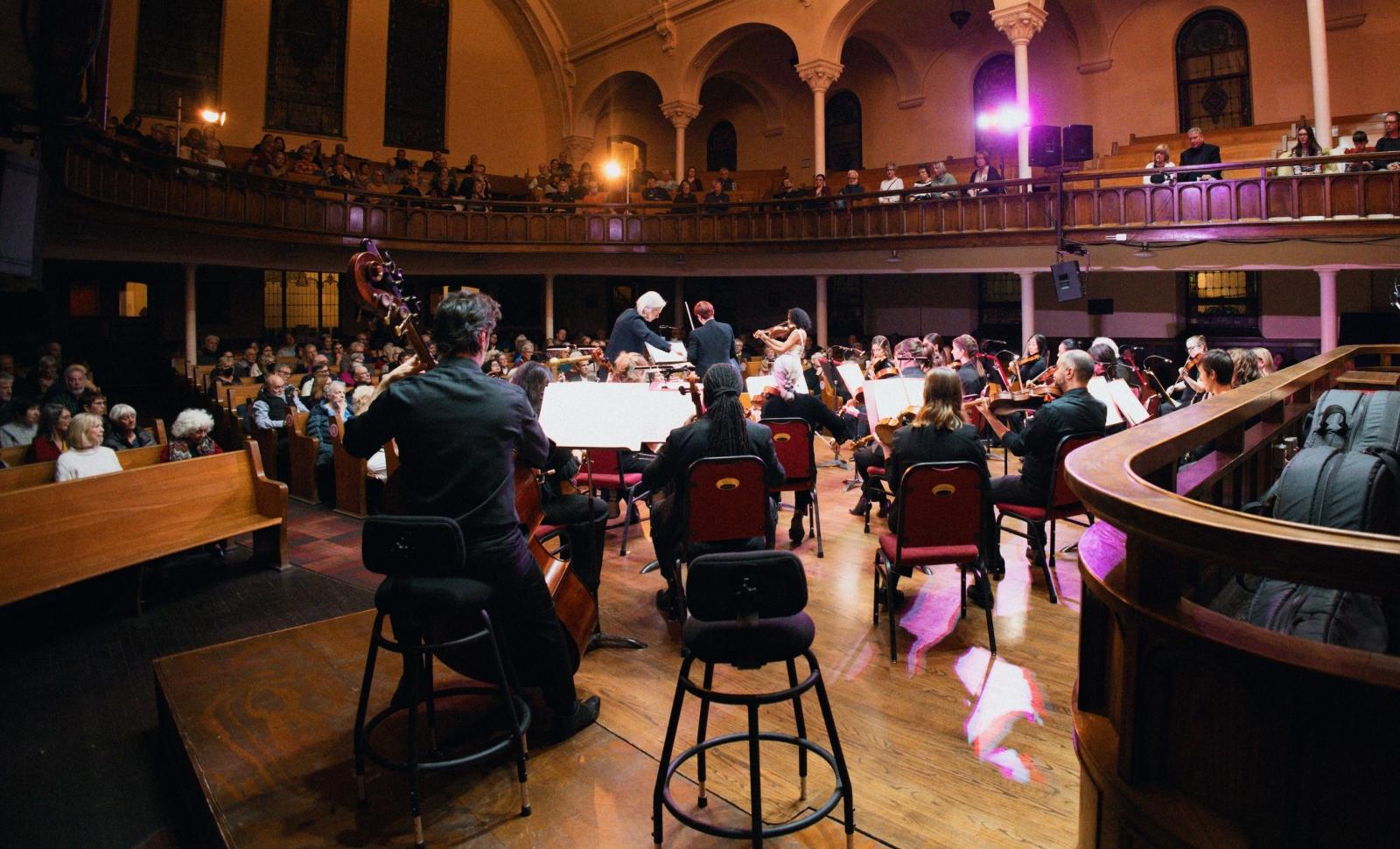 A chamber orchestra performs in a warmly lit concert hall with wooden balconies and arched architecture. Musicians seated in semicircles play string instruments while a conductor leads from the center. A solo violinist stands near the conductor. Audience members sit in pew-style seating on the main floor and upper balcony watching the performance.